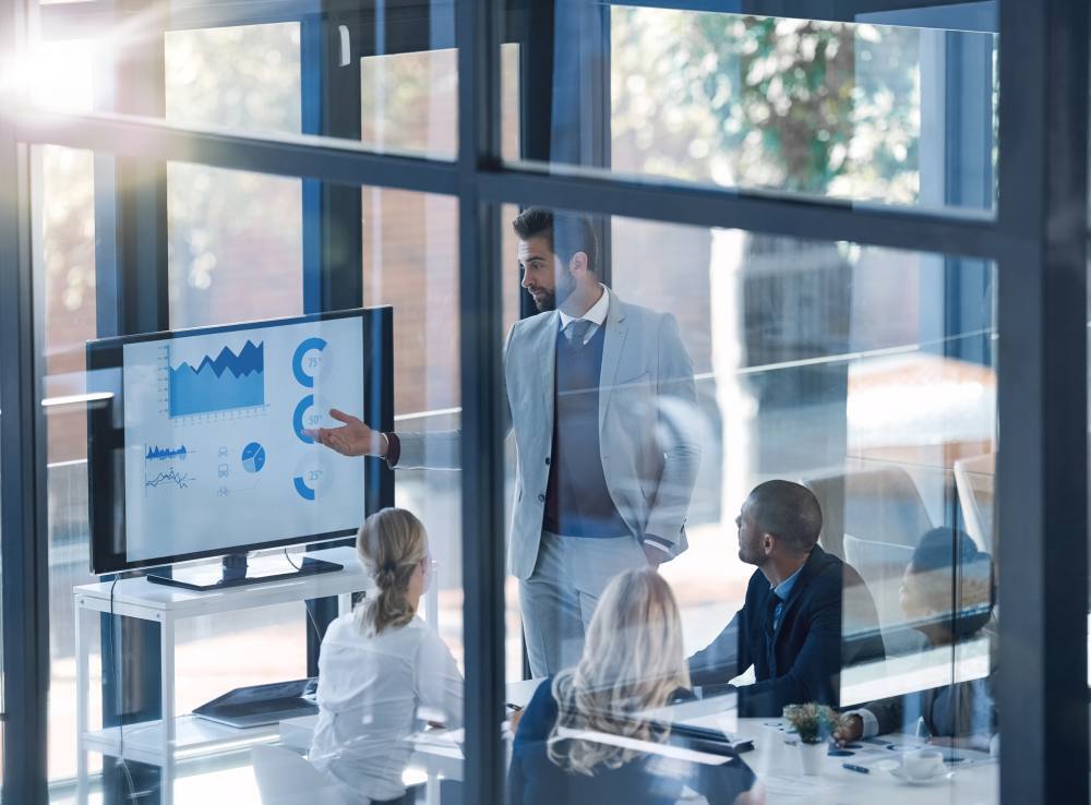 Un hombre con traje y corbata señalando gráficos coloridos en un monitor grande frente a dos colegas en una sala de reuniones corporativa.