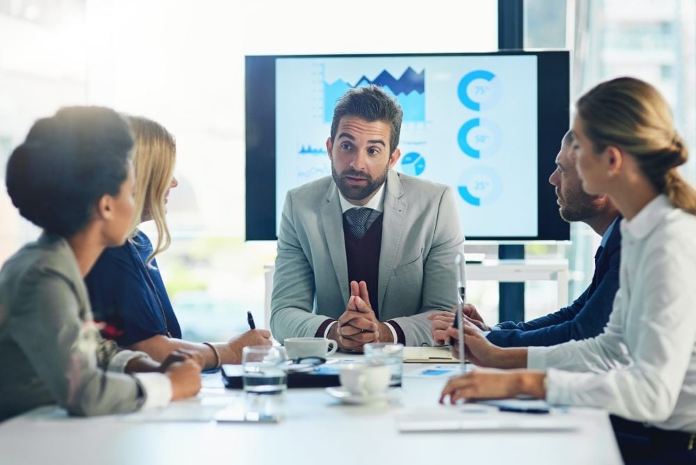 Un ejecutivo explicando gráficos en una pantalla a su equipo sentado en una sala de juntas, analizando la generación de demanda B2B