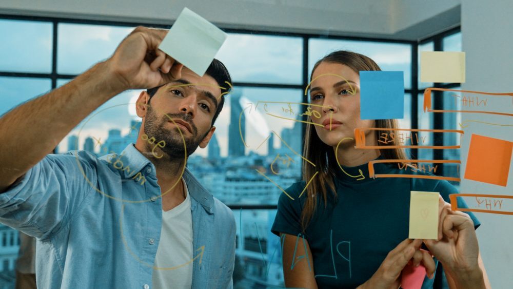 Dos personas planifican ideas pegando notas adhesivas en una pizarra de vidrio en una oficina.